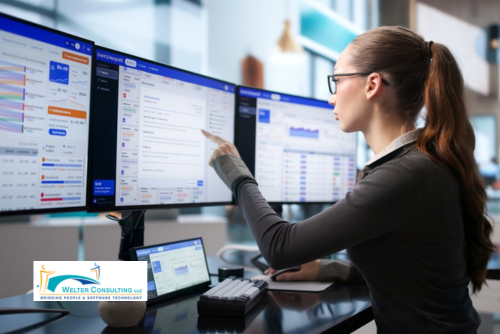 person sitting at desk in front of several computer monitors and pointing at one of them